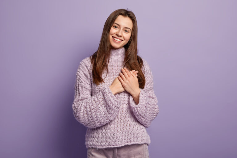 portrait happy young european woman keeps hands breast shows heart gesture expresses gratitude being thankful models against purple wall body language monochrome people devotion